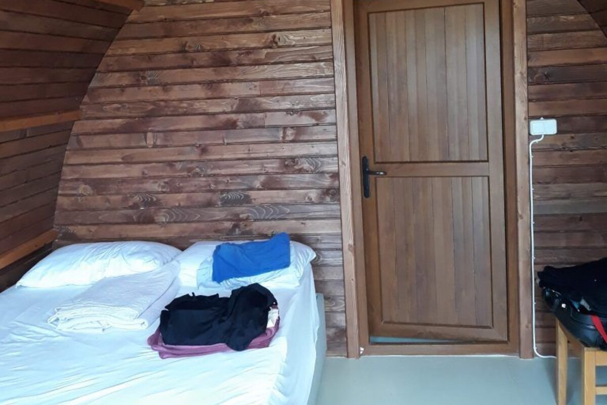 A small, cozy room with a sloped ceiling and wooden walls. It features a single bed with a white mattress and a blue blanket, a wooden door, and a small table with a chair.