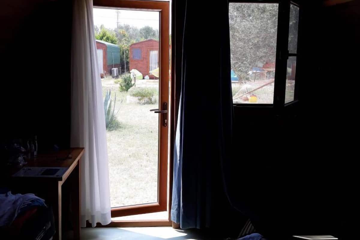 A room with a wooden ceiling, a bed, a desk, and a window overlooking a yard with a small building.