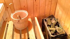 Una sala de sauna con un banco de madera, un cubo blanco y un recipiente con piedras.
