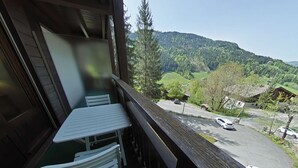A balcony with a table and chairs overlooking a valley with a road and parked cars. - Cozy 2-Room Apartment for 5 with Ski Storage and Garage in Le Grand-Bornand (Le Grand-Bornand)
