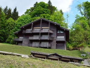 A modern multi-story building with balconies, surrounded by lush greenery and trees.
