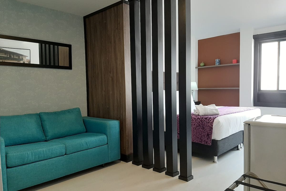 A modern hotel room with a teal sofa, a bed with a purple and white bedspread, a wooden partition, and a window with a view.