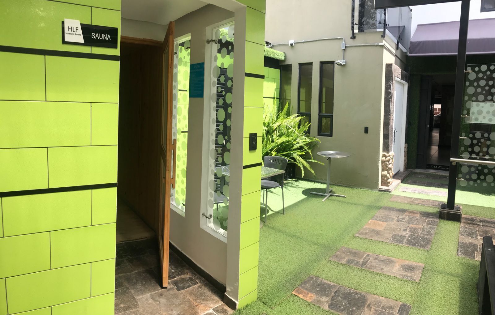 Área de spa com parede revestida de azulejos verdes, placa de sauna, janela com padrão de poás, uma pequena mesa e uma cadeira.