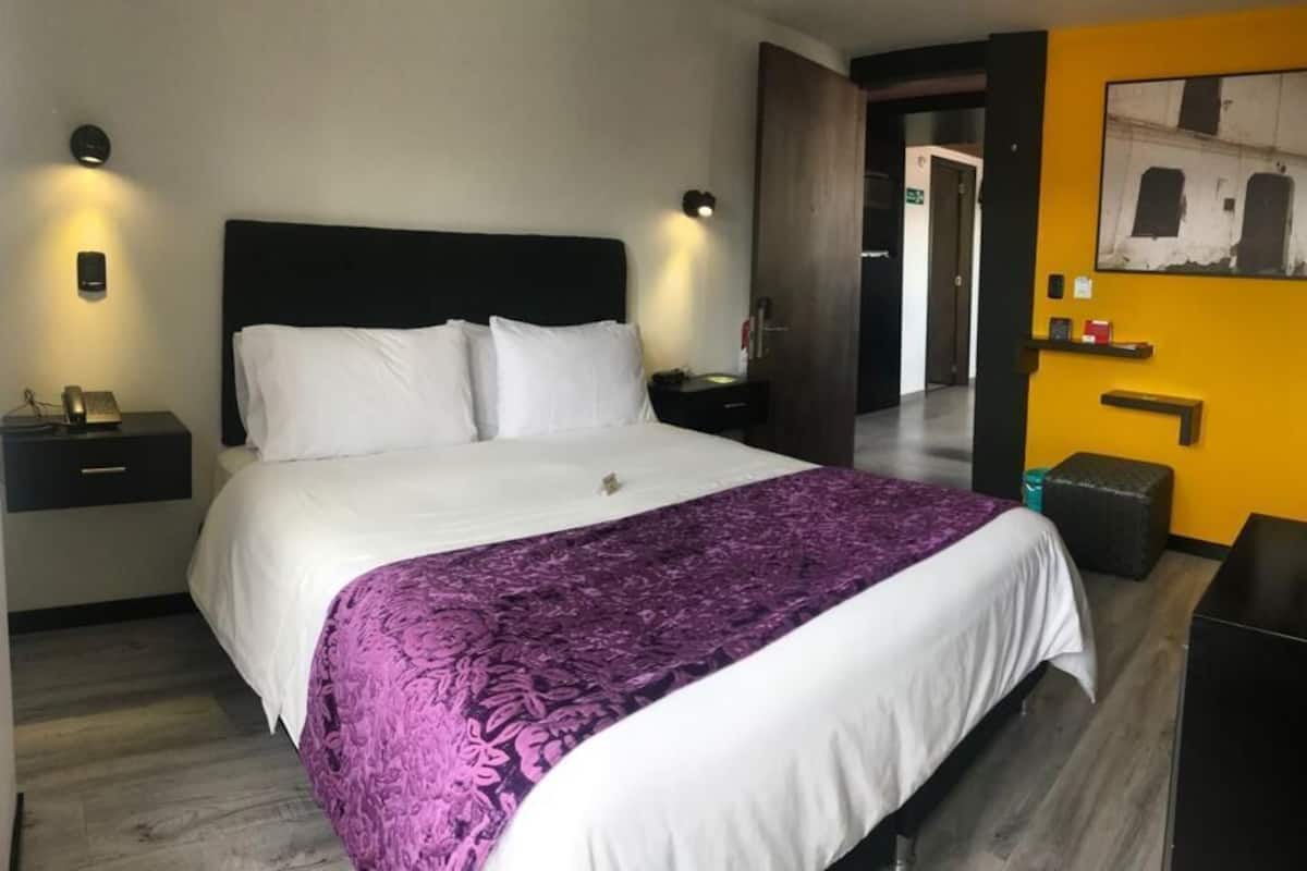 A hotel room with a large bed, a nightstand with a phone, a wall-mounted light, and a door leading to another room.