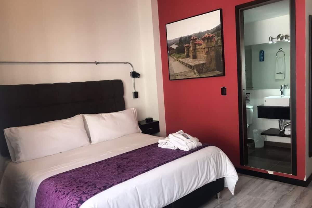 A hotel room with a bed, a purple blanket, a white pillow, a nightstand, a wall-mounted lamp, a framed picture, a sliding glass door to the bathroom, and a red wall.