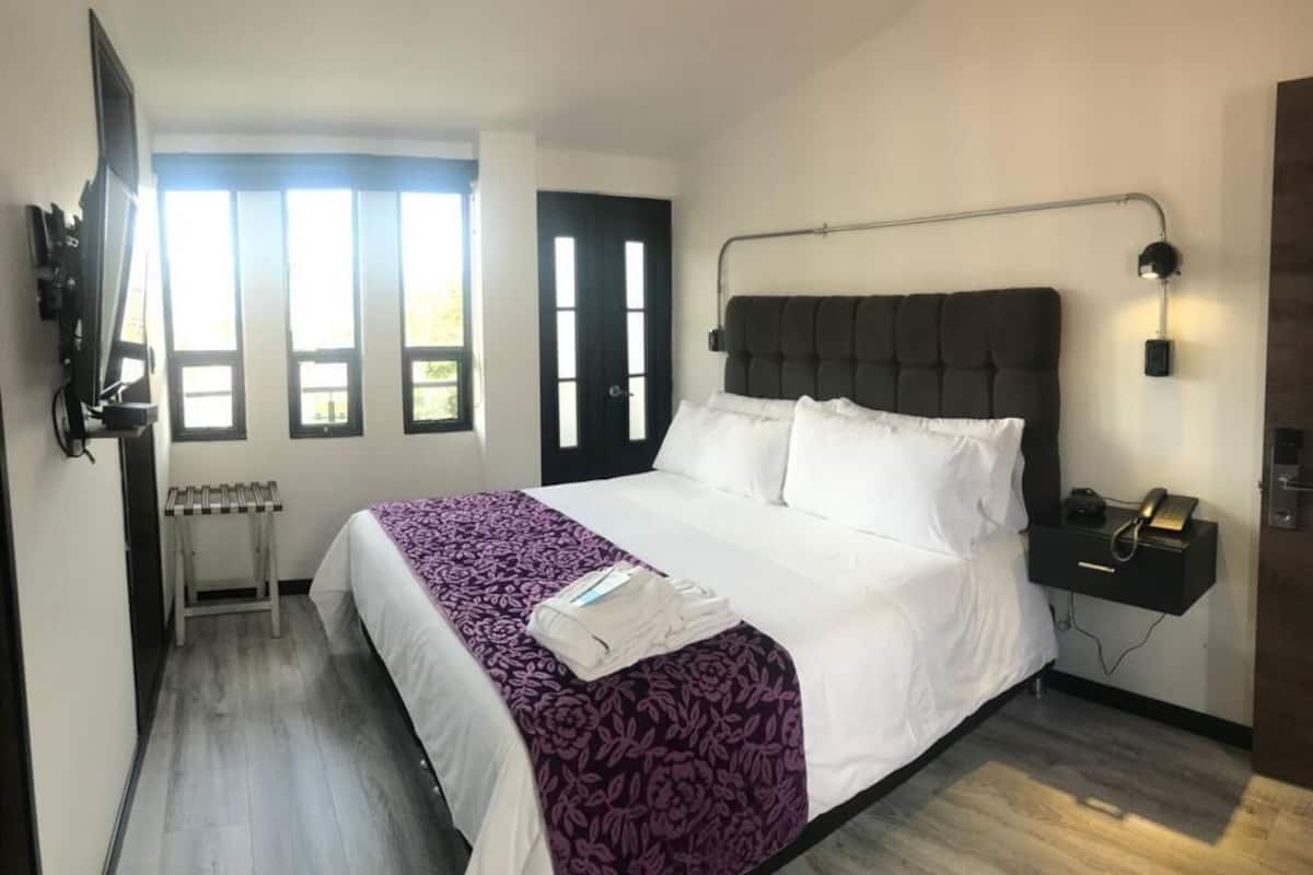A hotel room with a large bed, a bedside table with a phone, a wall-mounted lamp, and a window with curtains.