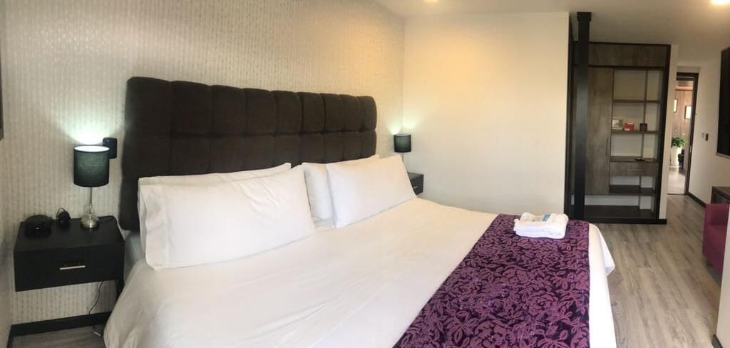 Ett hotellrum med en stor säng, två sängbord med lampor, en mörk sänggavel och en lila pläd.