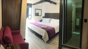 Habitación de hotel con cama, sofá rojo, puerta de madera y cabina de ducha de vidrio.