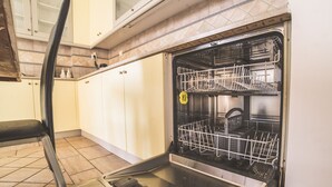 Un lave-vaisselle ouvert dans une cuisine dont le sol est carrelé et les meubles de rangement sont blancs.