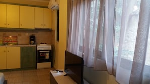 A small kitchen with yellow cabinets, a stove, and a coffee maker. There is a window with sheer curtains. - Central Studio (Patras)