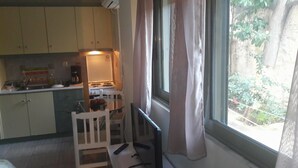 A compact kitchen with a stove, sink, and cabinets. There is a dining table with chairs and a window with curtains. - Central Studio (Patras)