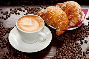 A cup of coffee with latte art on a saucer, two croissants, and scattered coffee beans on a wooden surface. - Riviera Luxury Suites B&B (Naples)