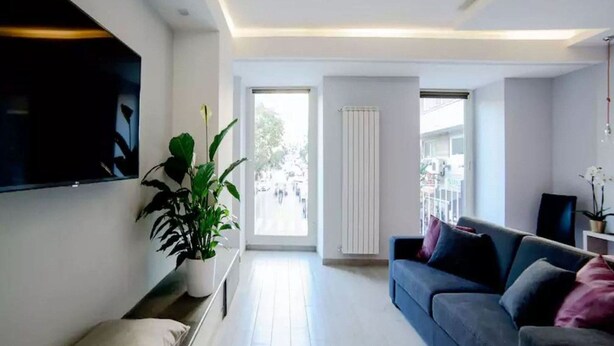 A modern living room with a flat-screen TV mounted on the wall, a dark-colored sofa with cushions, a potted plant, and a sliding glass door leading to an outdoor area. - Riviera Luxury Suites B&B (Naples)