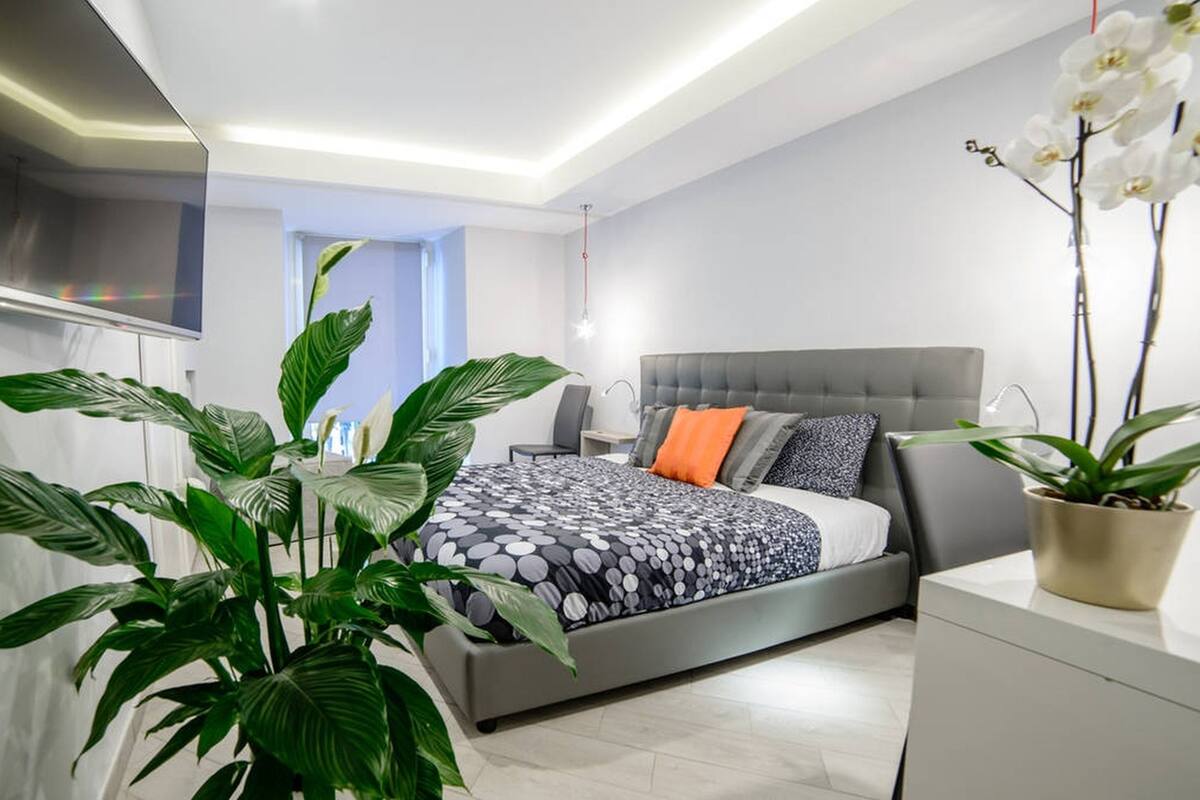 Une chambre moderne avec un lit gris, des murs blancs et des plantes vertes.
