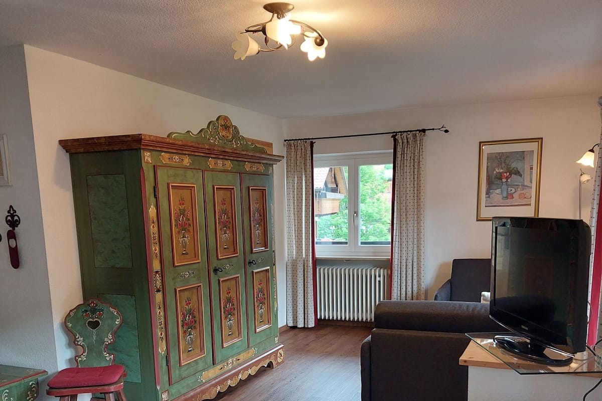 Quarto com cama, um armário verde com motivos florais, televisão sobre um suporte e janela com cortinas.