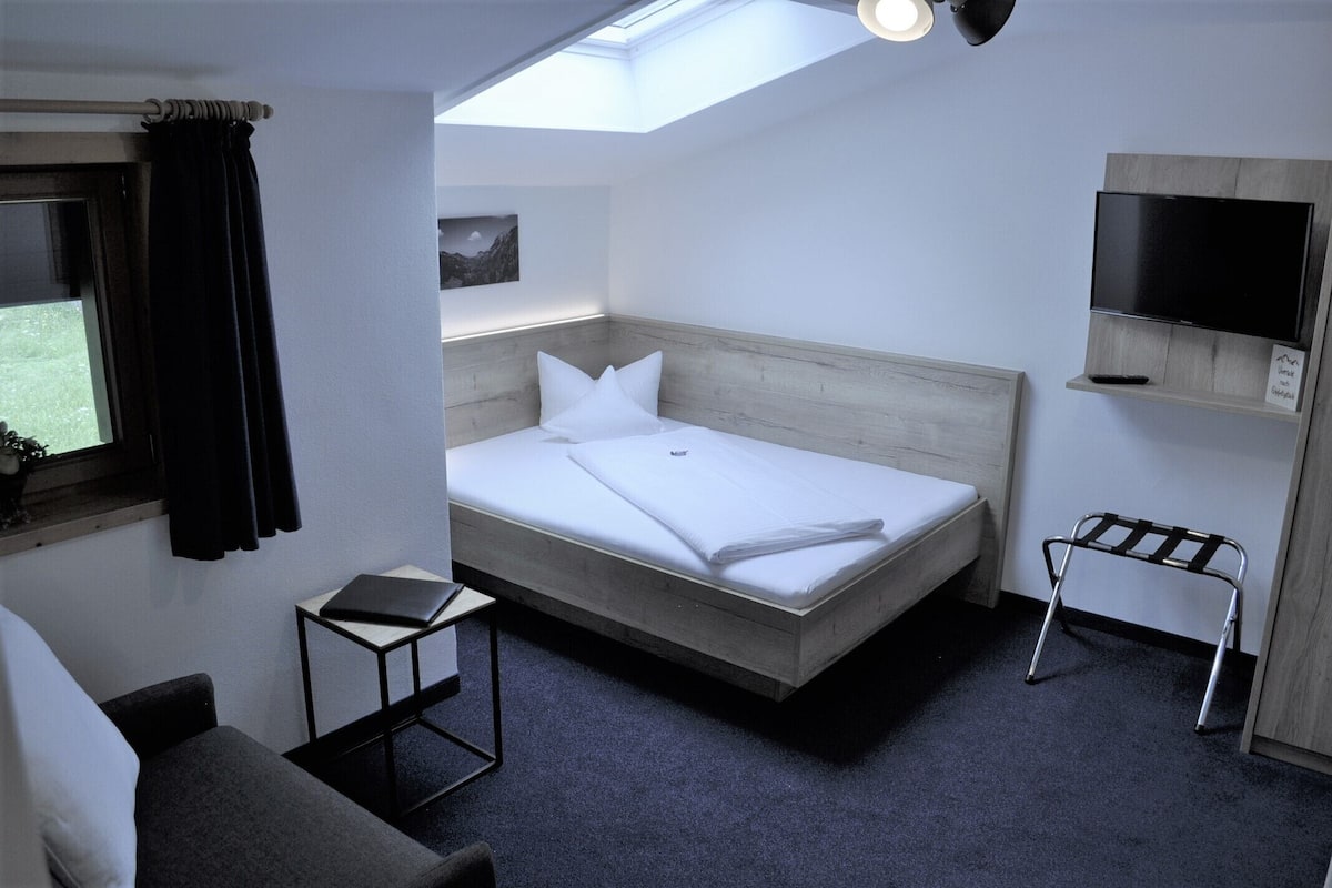 Quarto de hotel moderno com claraboia, cama, mesinha, televisão e uma cadeira.