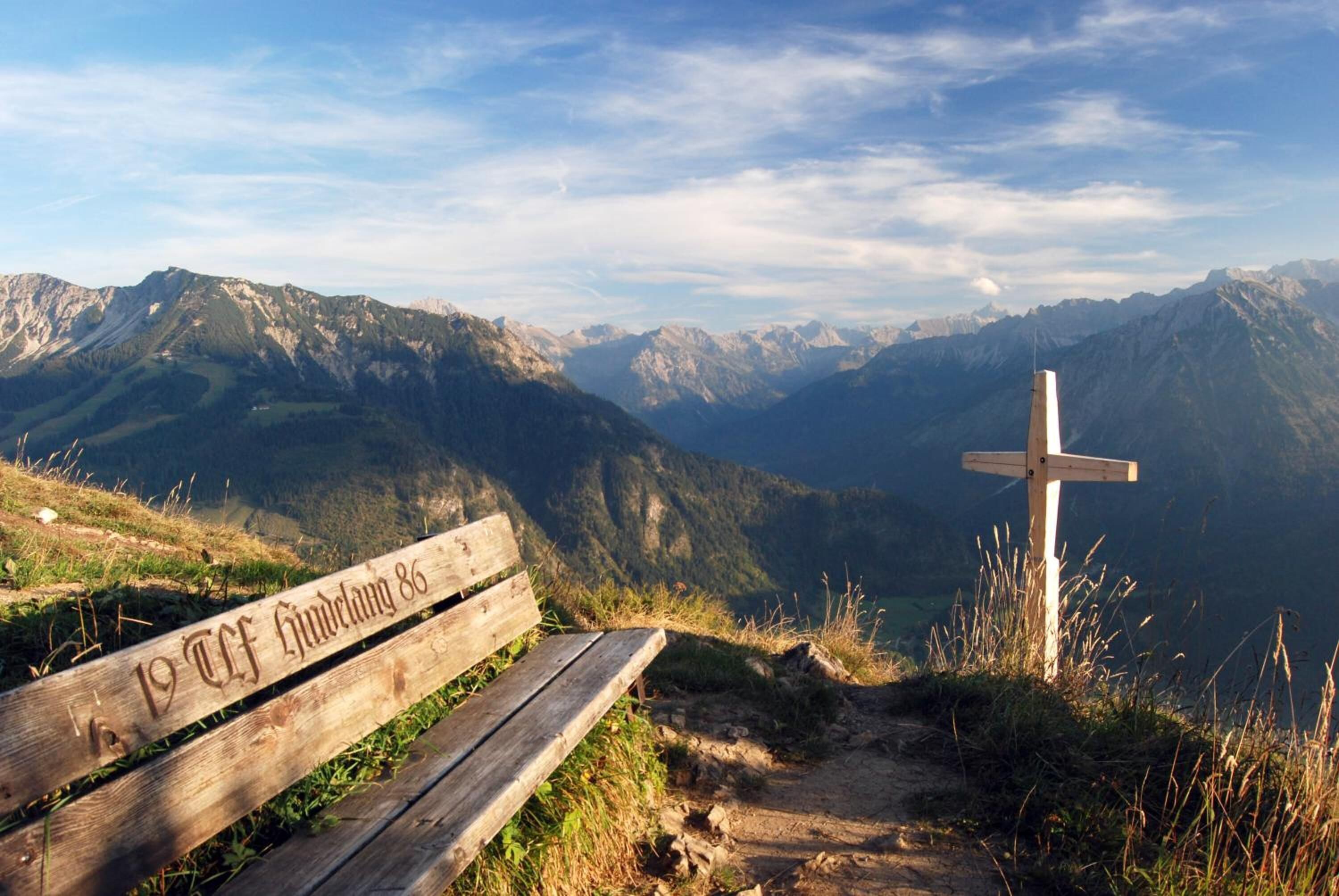 A wooden bench with "19 CF Churwald 86" engraved on it, overlooking a mountainous landscape with a wooden cross in the foreground.
