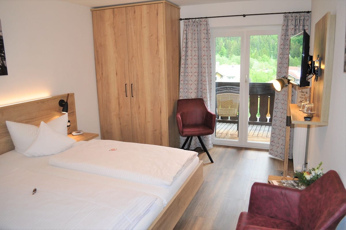 Quarto com cama, guarda-roupa, cadeira, escrivaninha com abajur e vista através de uma porta de vidro.