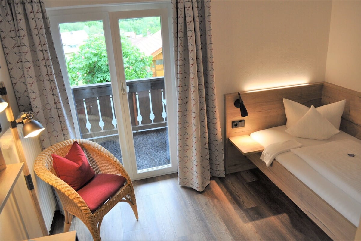 Quarto de hotel com uma cama, uma cadeira de vime com almofada vermelha, uma mesa de cabeceira com abajur e vista para o verde através de uma porta de vidro.