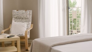 A hotel room with a bed, a wooden chair with a leaf-patterned cushion, a small table with a remote control, and a window with a view of greenery. - Casa Mia Apartments (Agios Nikolaos)