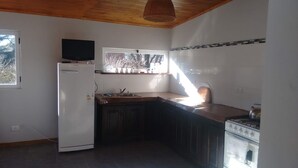 A compact kitchen with a refrigerator, a stove, a window with a view, and a television on the wall. - Casita del Sauce (Sierra de la Ventana)