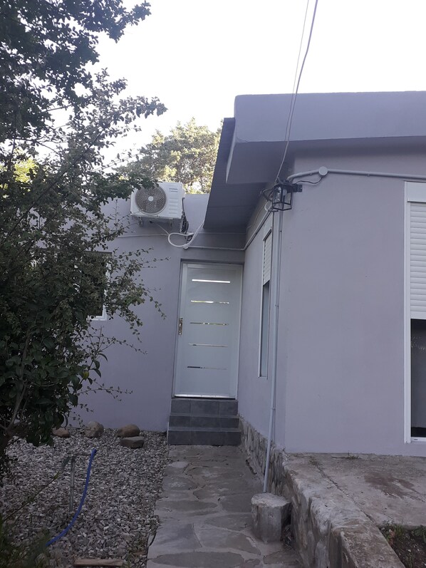 A narrow stone pathway leading to a door with a small step, a white wall with a window, and an air conditioning unit mounted on the wall.