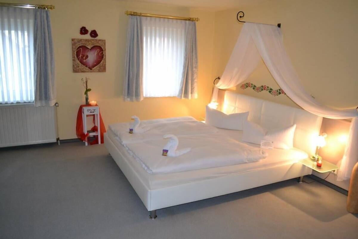 A bedroom with a bed, a bedside table with a lamp, a wall decoration, and a window with curtains.