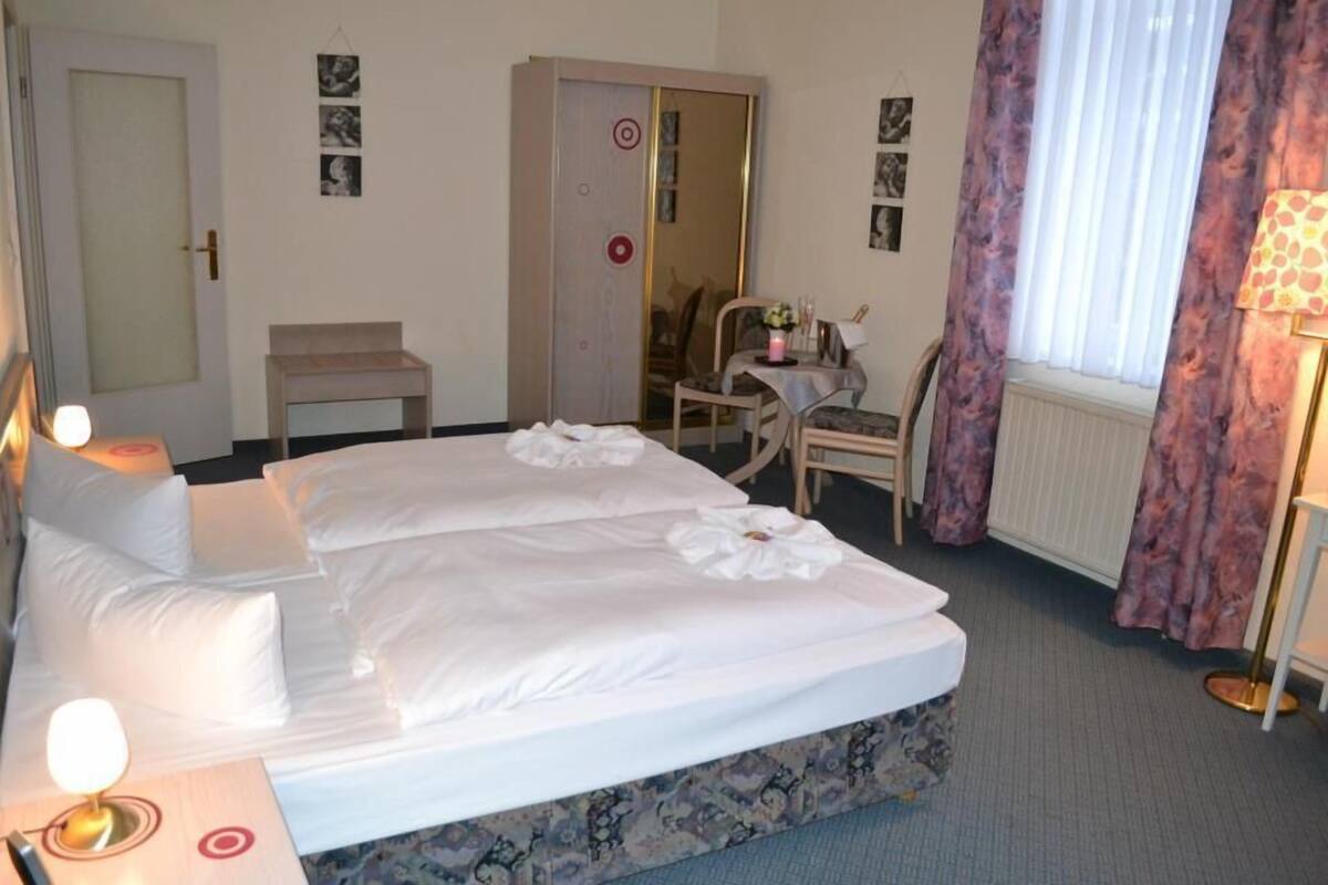 A hotel room with a bed, bedside lamps, a chair, a small table, and a window with curtains.