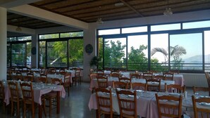 Uma área de refeições com mesas preparadas para hóspedes, grandes janelas com vista para o verde e montanhas, e um teto com vigas aparentes.