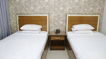 Habitación de hotel con dos camas individuales, cabeceros de madera, ropa de cama blanca, una mesita de noche con teléfono y un papel tapiz estampado con motivos florales.