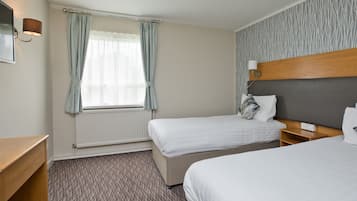 A hotel room with two beds, a wooden headboard, a small bedside table, a nightstand, a lamp, and a window with curtains.