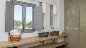A bathroom with a large window, two mirrors, a wooden vanity, and a stone wall. - Saint Apostles (Santorini)
