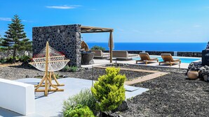A rooftop pool area with lounge chairs, a hanging chair, and a stone wall. - Saint Apostles (Santorini)