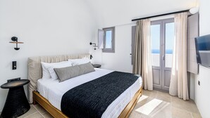 A bedroom with a bed, bedside tables, a TV, and a window with curtains. - Saint Apostles (Santorini)