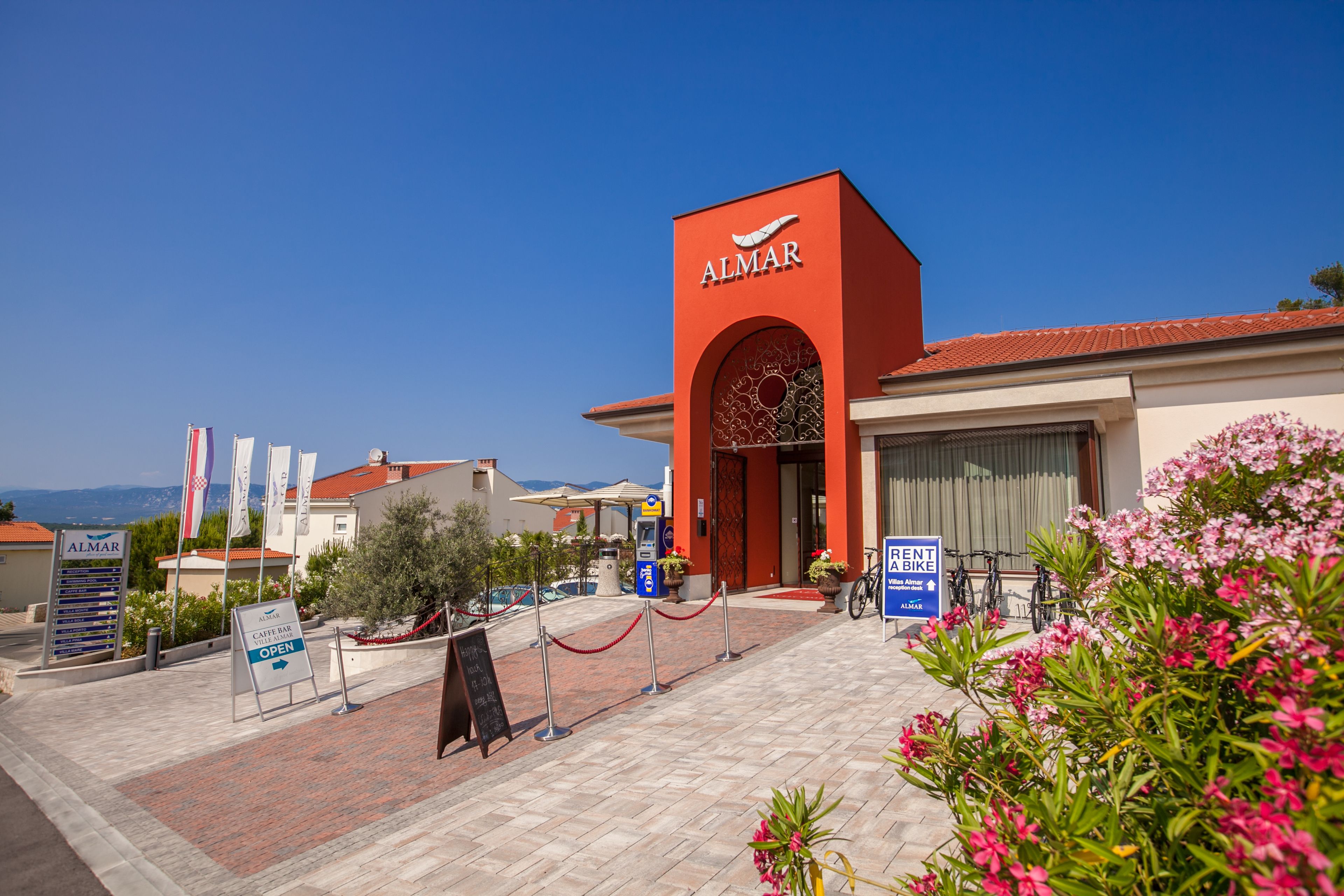 Entrada de hotel com fachada vermelha, placa indicando café bar e serviço de aluguel de bicicletas.