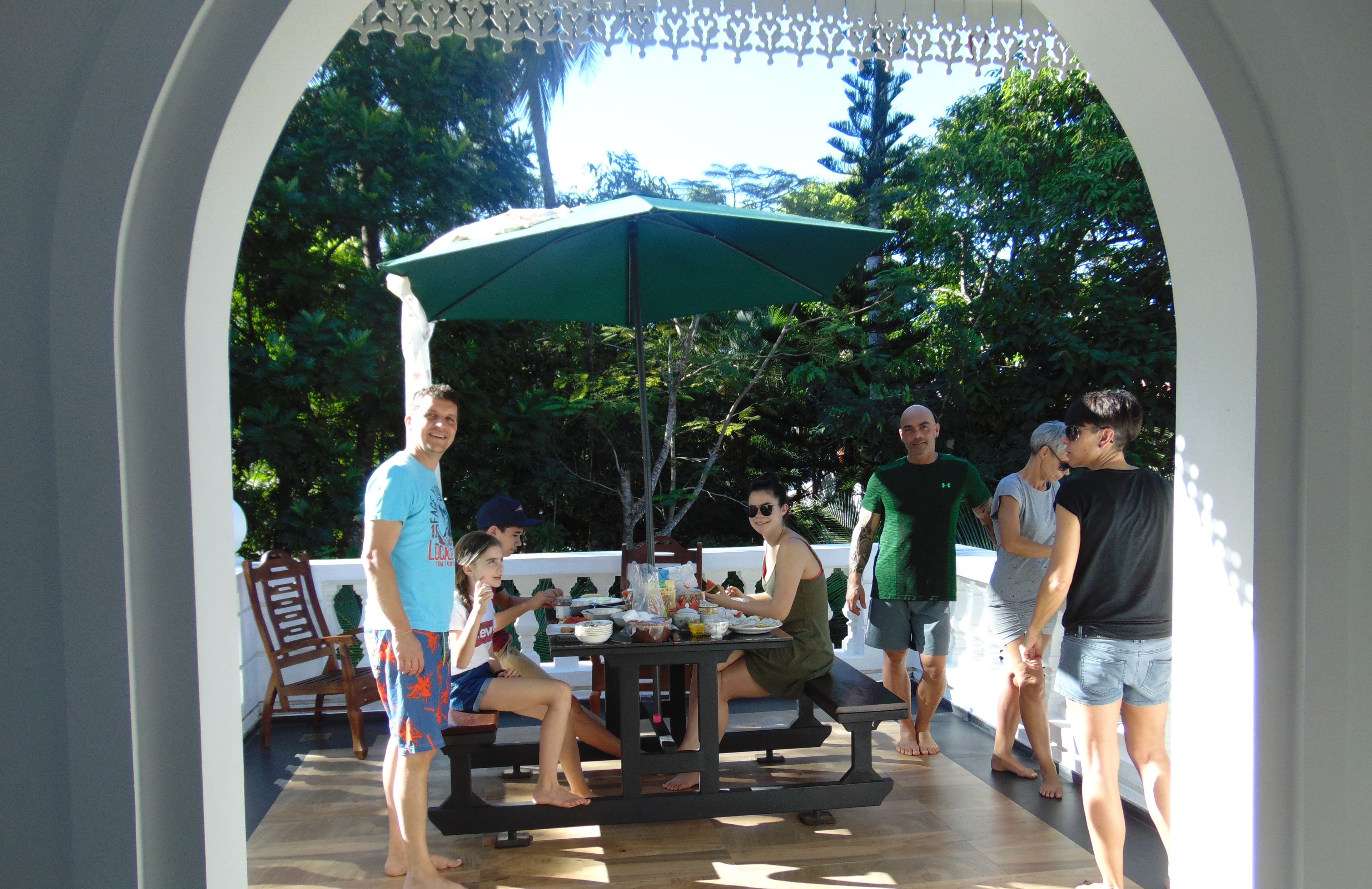 A group of people dining at an outdoor table under a green umbrella, surrounded by a white fence and lush greenery.