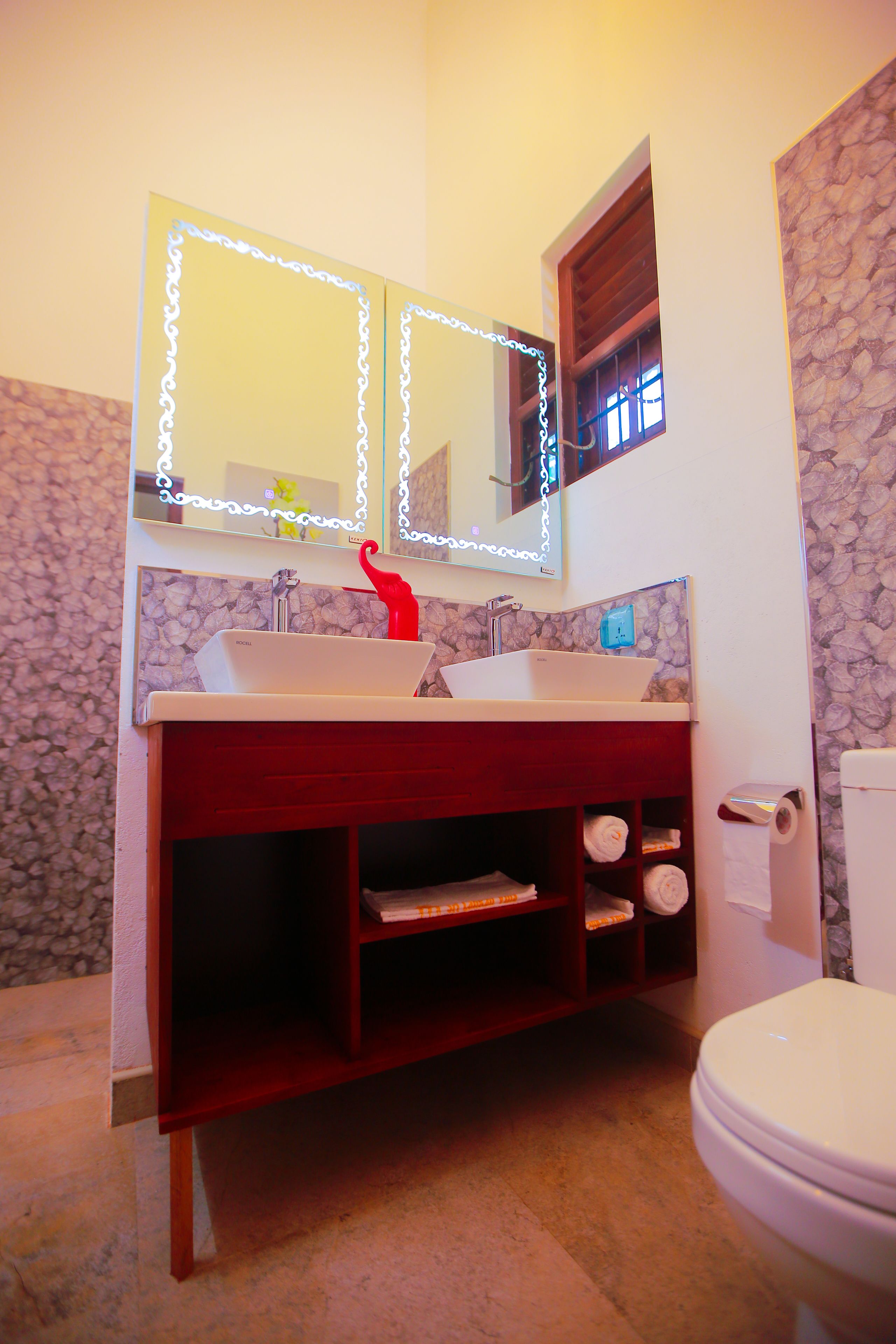 Ein Badezimmer mit einem Doppelwaschbecken, einem roten Dekoelement, einem Spiegel mit Beleuchtung, einem Fenster und einer Toilette.
