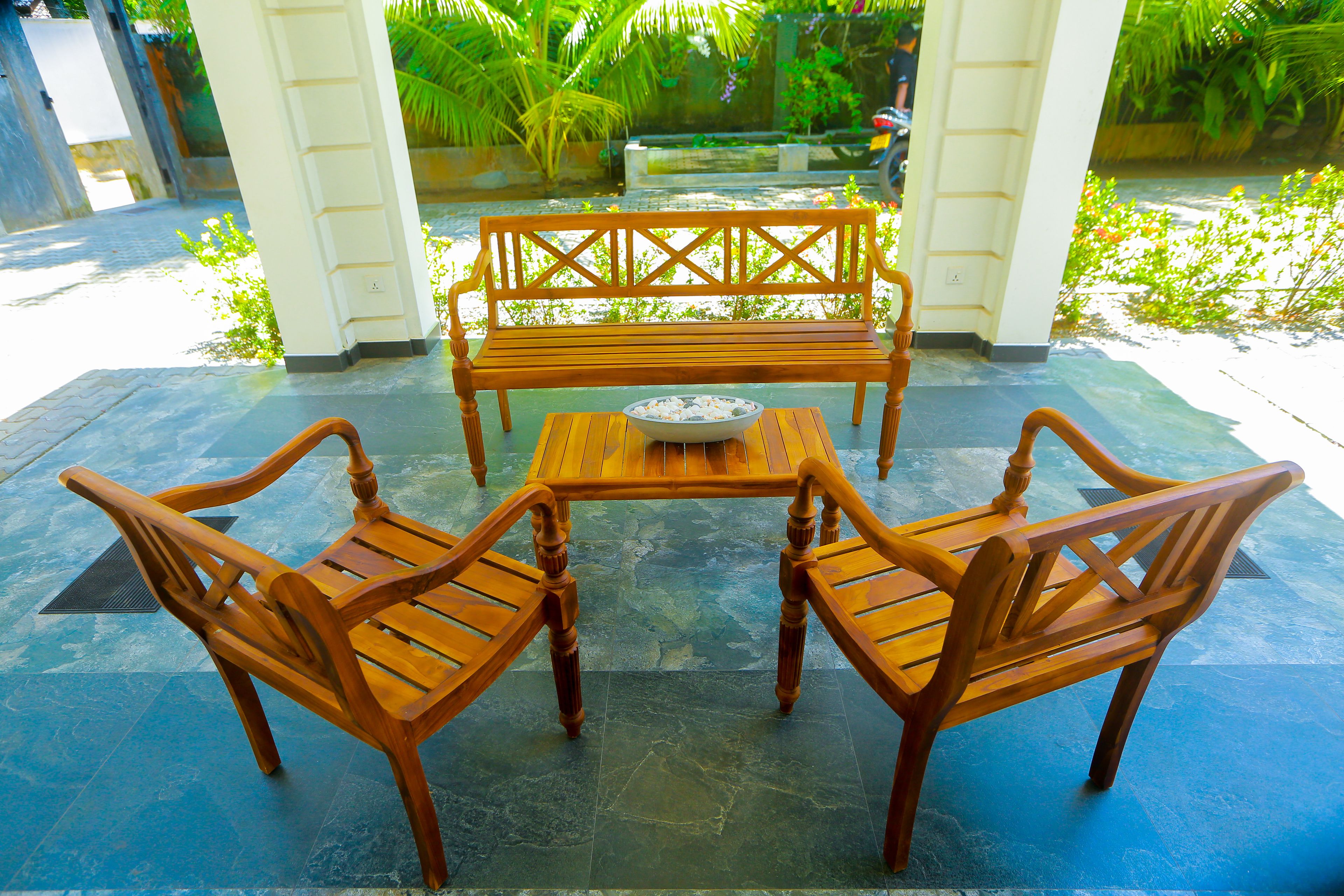 A wooden bench and table set on a tiled patio with a view of greenery and a parked motorcycle in the background.