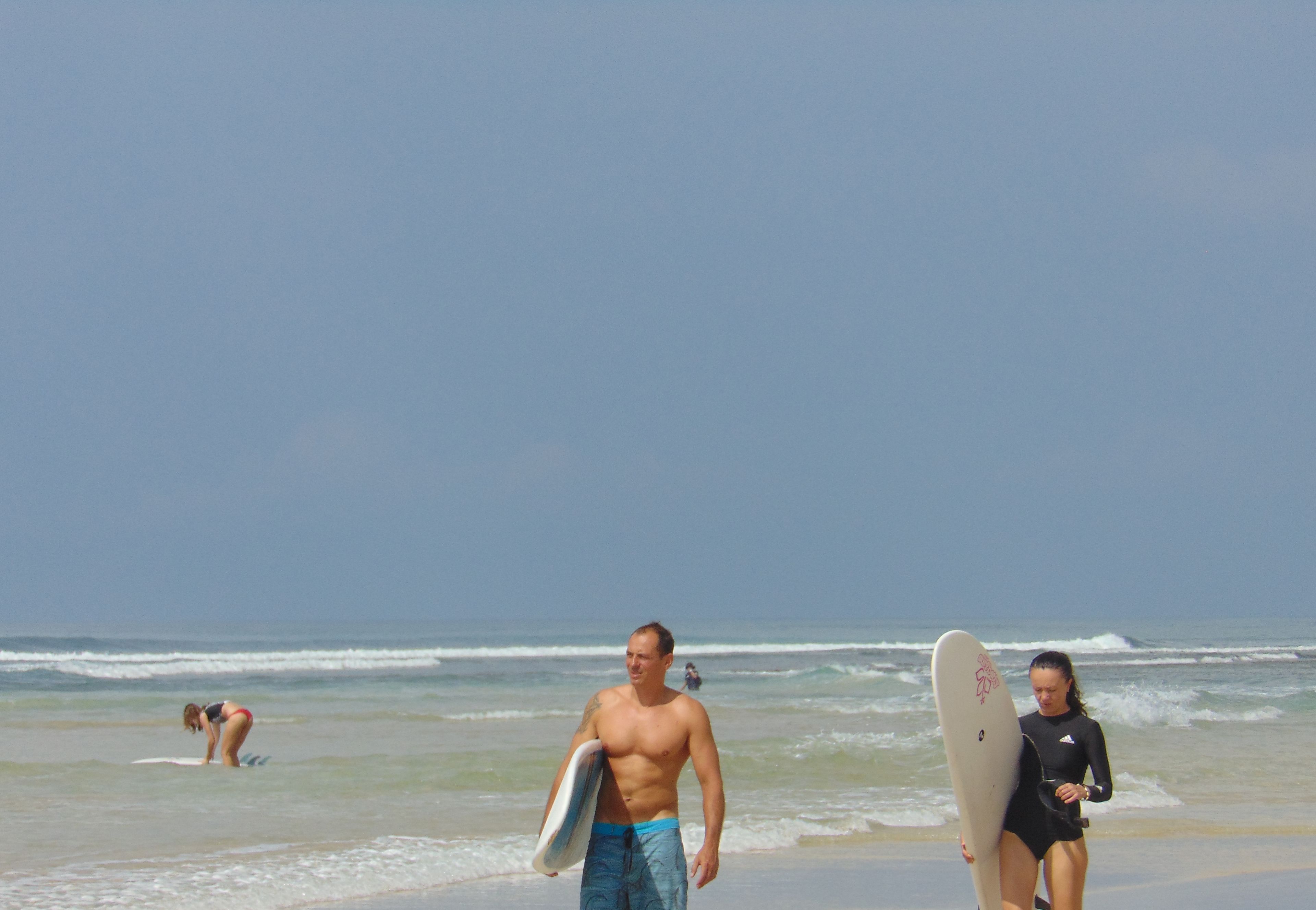Escena de playa con un hombre y una mujer caminando con tablas de surf.