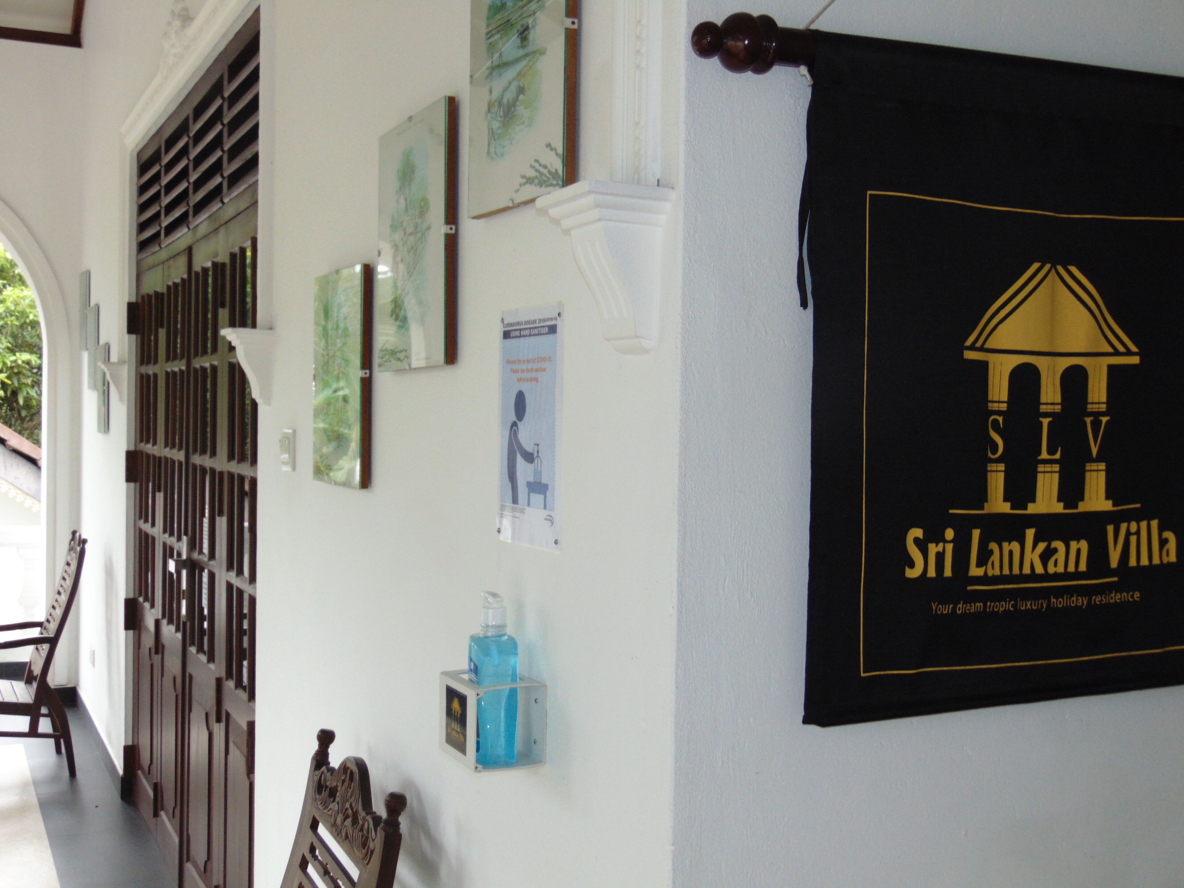 Un pasillo con obras de arte enmarcadas, un dispensador de gel hidroalcohólico y un cartel para Sri Lankan Villa.