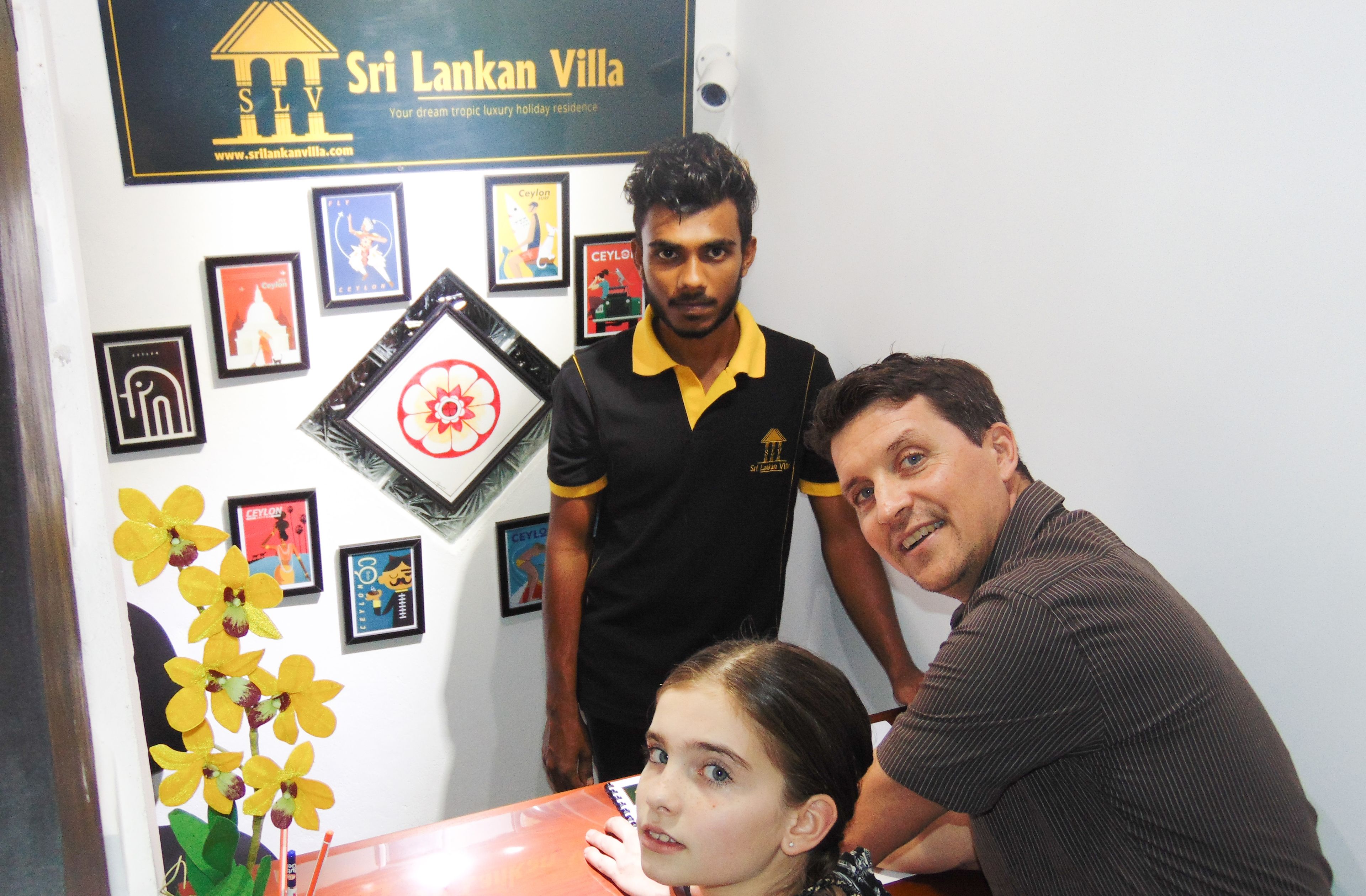 A reception area with framed pictures on the wall, a desk with a flower arrangement, and a sign for Sri Lankan Villa.