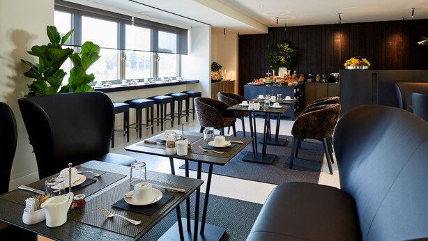 A modern dining area with tables set for breakfast, large windows, and a kitchen in the background. - Delparco Hotel (Buttrio)