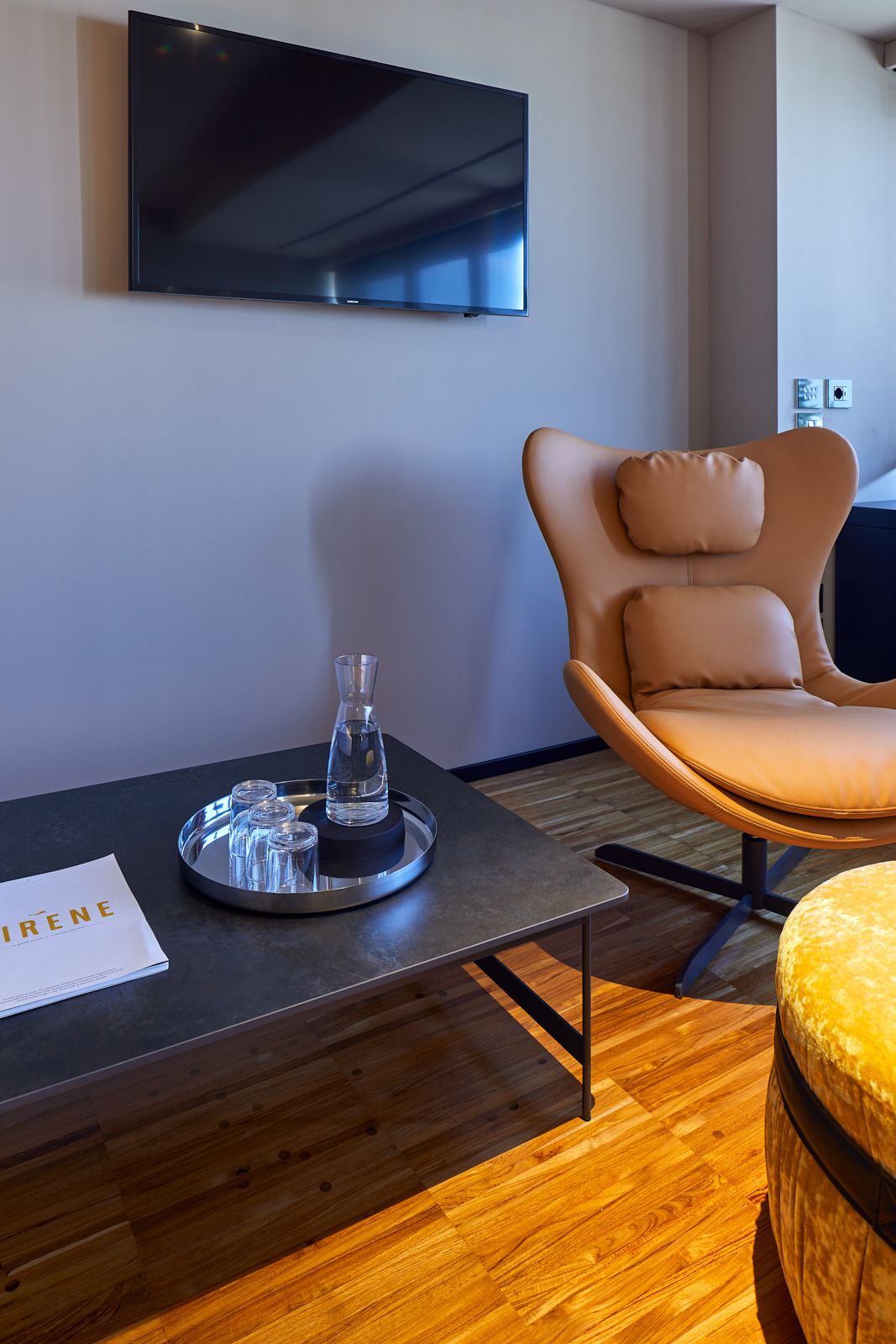 A modern room with a flat-screen TV mounted on the wall, a leather chair, a wooden floor, and a small table with a tray holding glasses and a bottle.
