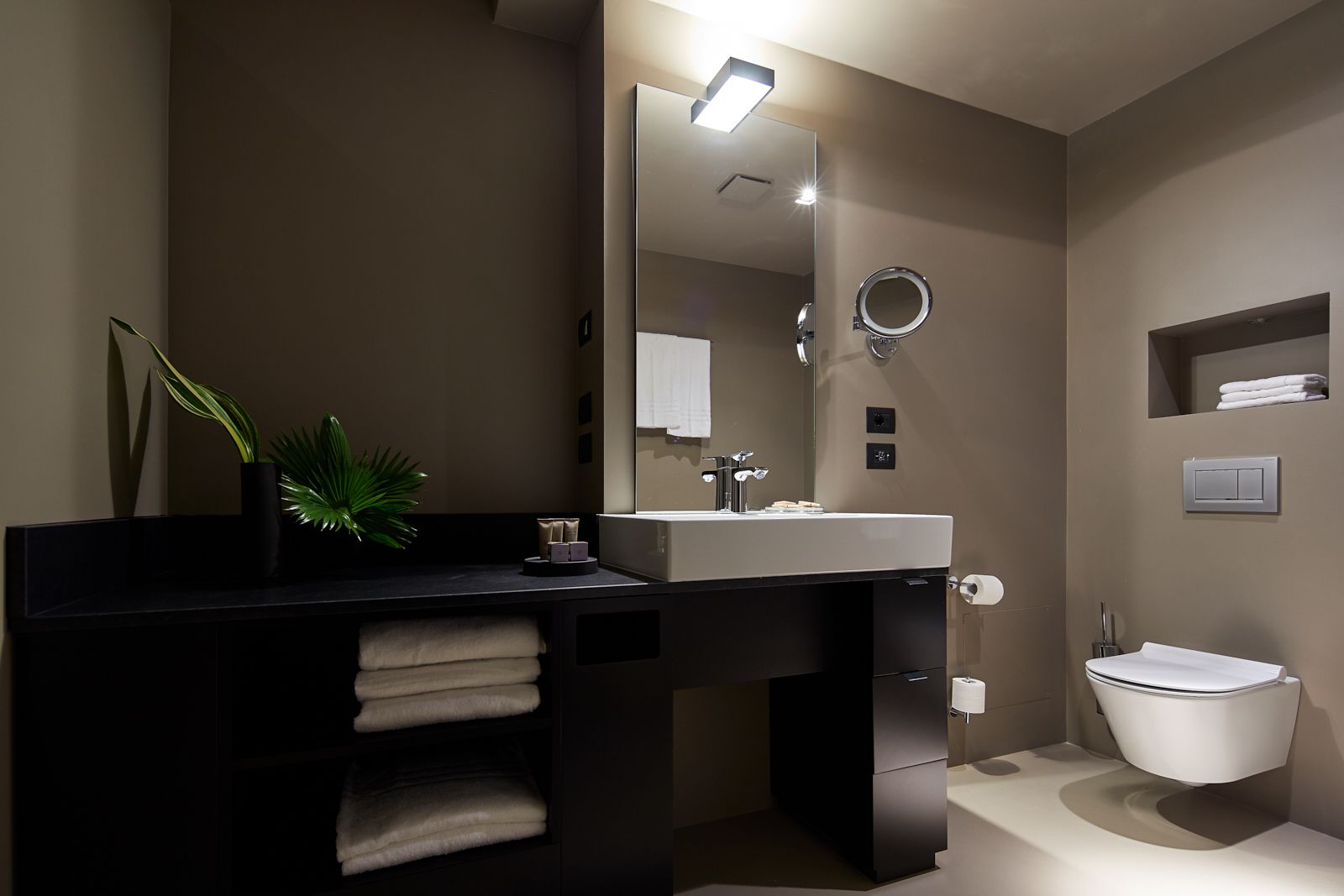 A modern bathroom with a black vanity, white sink, and a large mirror. There is a plant on the vanity and a towel rack below it.