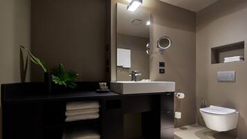 A modern bathroom with a black vanity, white sink, and a large mirror. There is a plant on the vanity and a towel rack below it.