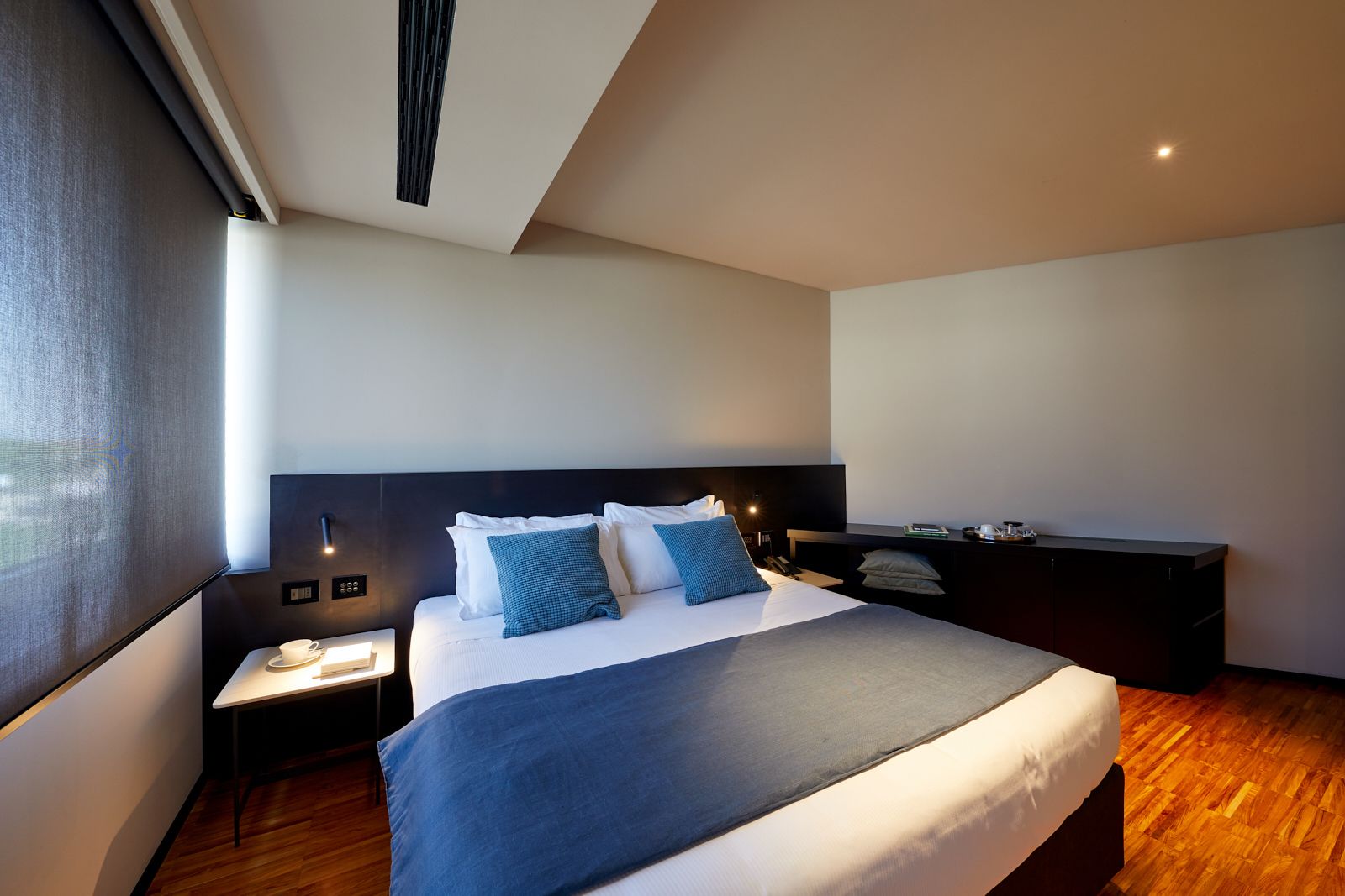 A modern hotel room with a large bed, a bedside table with a lamp, a small desk, and a large window.