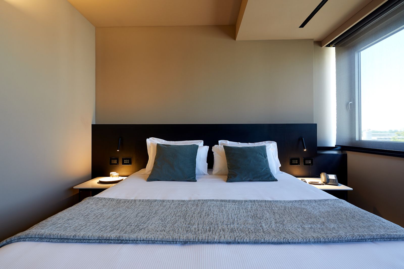 A hotel room with a large bed, two bedside tables with lamps, a window with a view, and a modern headboard.