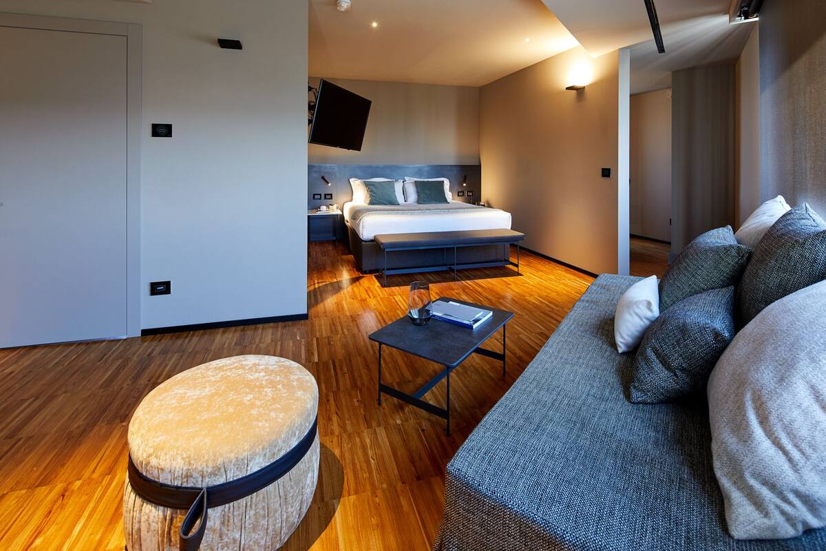 A modern hotel room with a large bed, a sofa, a small table, and a round ottoman.