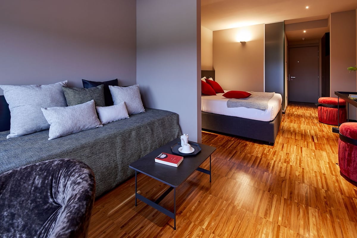 A modern hotel room with a sofa, a small table with a book, a bed with red pillows, and a wooden floor.