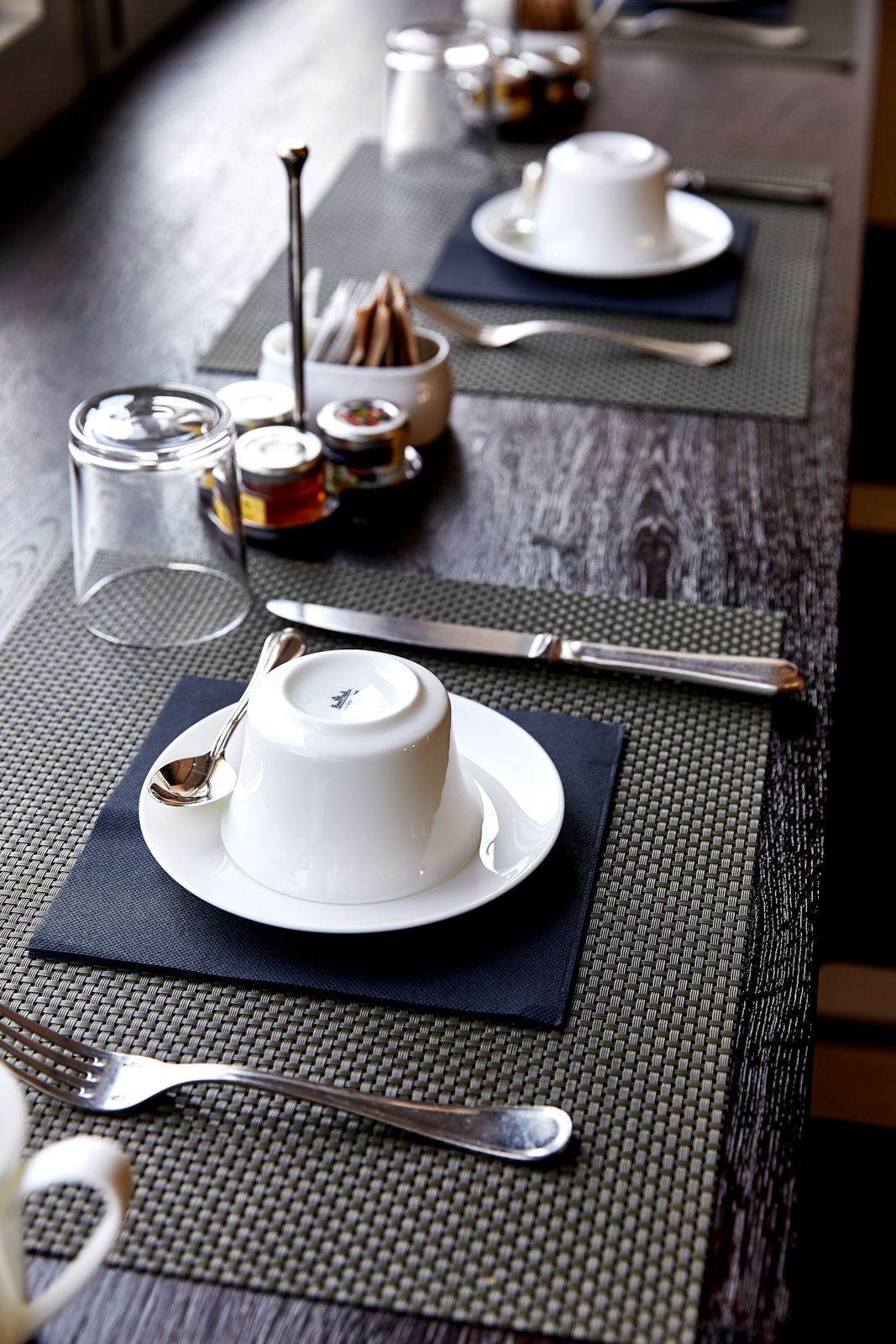 Un tavolo da pranzo apparecchiato con piatti bianchi, posate e una tazza, con un sottopiatti in tessuto.